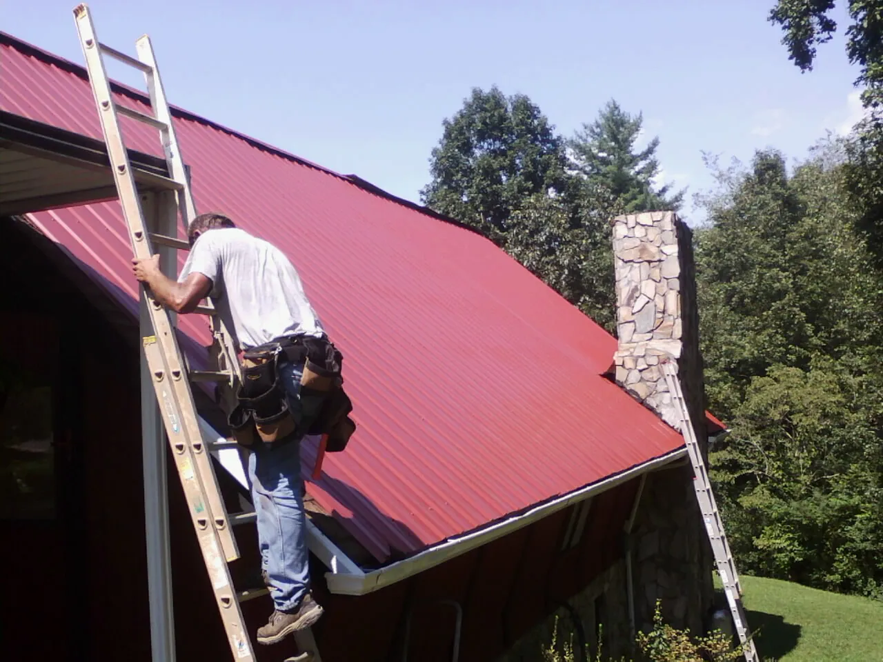Roofer on a ladder beside a red metal roof and stone chimney — Soffit & Fascia Repair by Roofing Brothers in Prairie Village